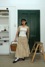 Woman in a white top and beige skirt standing in a room with a green door and wooden stool.